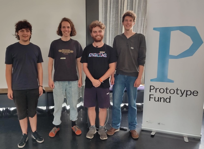 A group photo of the four team members in front of a banner with the prototype fund logo.